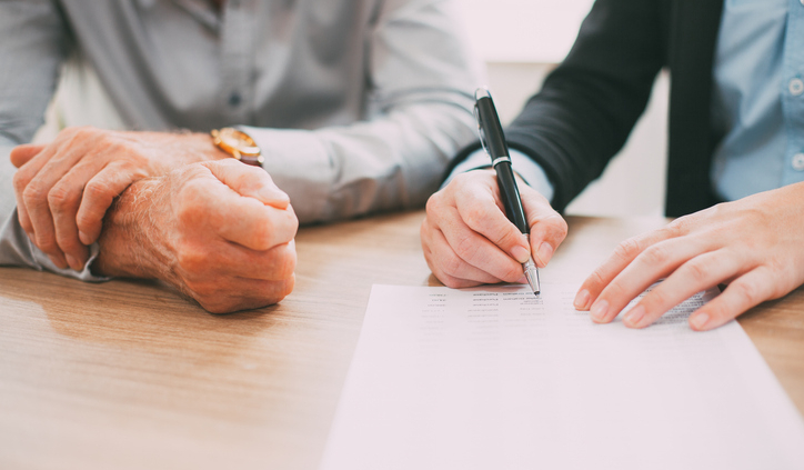 Two people sit at a desk with one of them signing a letter representing the letter of intent to buy a business.