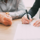 Two people sit at a desk with one of them signing a letter representing the letter of intent to buy a business.